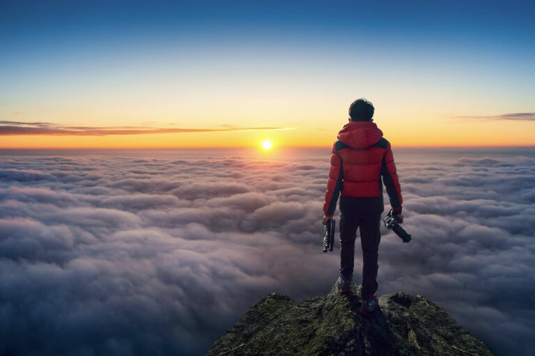 Человек стоит один на вершине горы над облаками, глядя на восход солнца